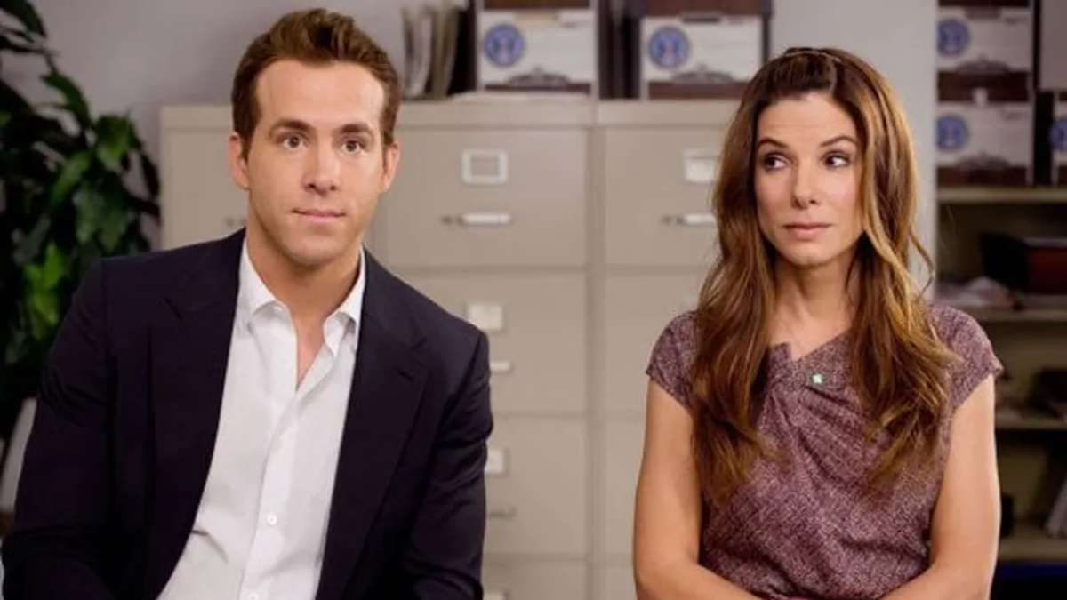 Ryan Reynolds e Sandra Bullock sentados lado a lado em cena de A Proposta