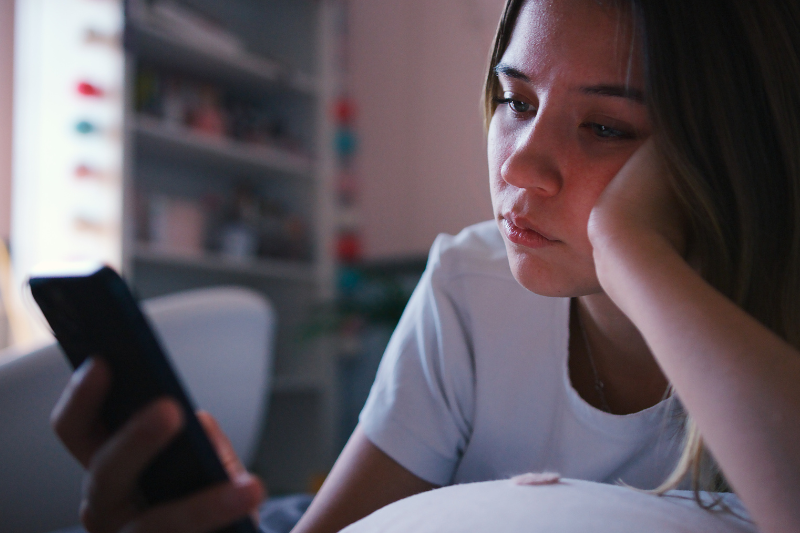 Uma menina adolescente deitada entediada com uma mão no rosto, enquanto a outra mão mexe no celular