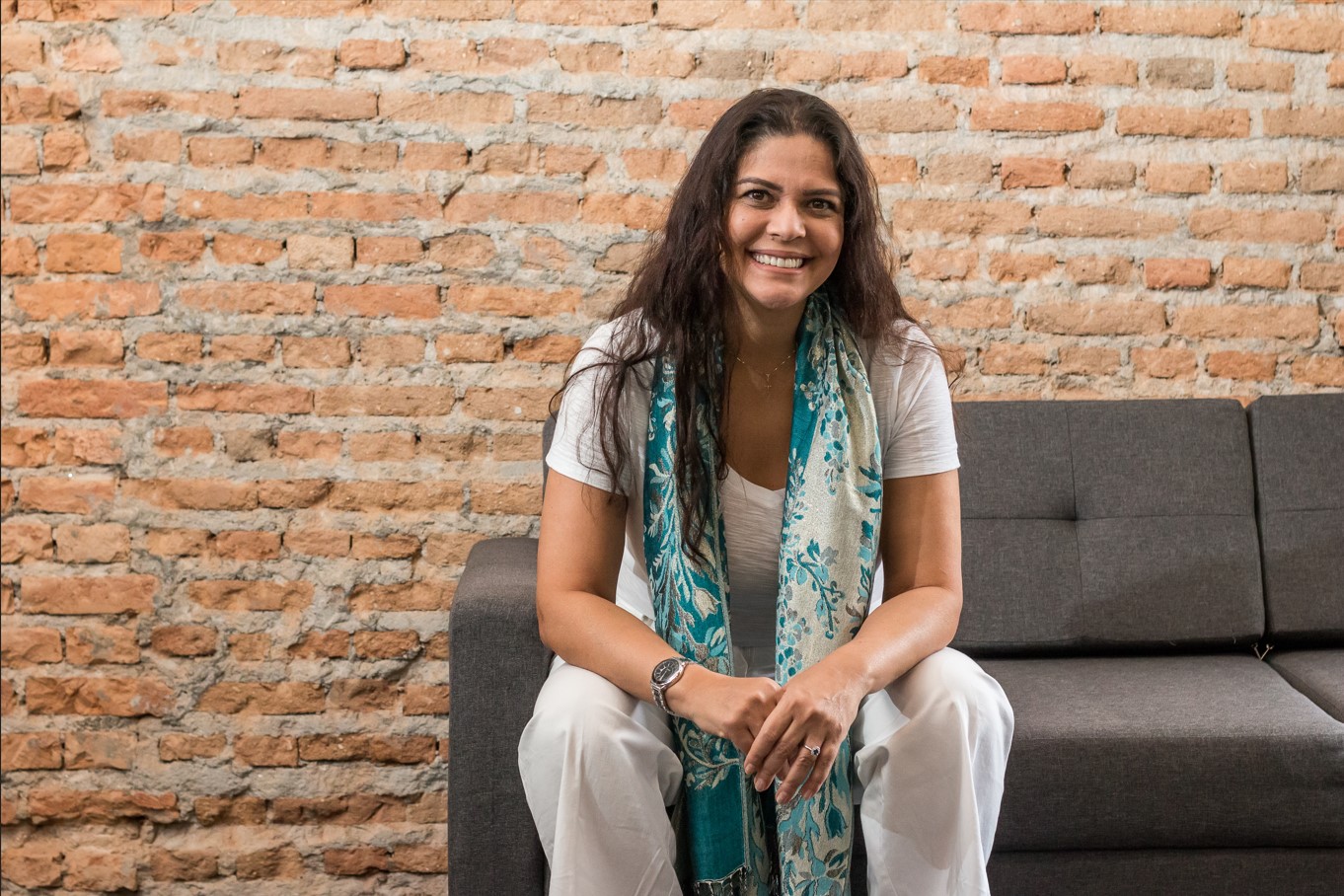 Chris Sabbag sentada em um sofá, sorrindo e com echarpe no pescoço caindo a frente de seu corpo