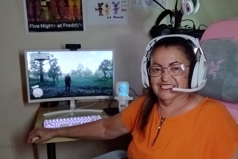  Texto Alternativo (Alt Text) para a Imagem 👩‍🦳🎮 Mulher idosa sentada em cadeira gamer, sorrindo e usando headset branco enquanto joga em seu PC