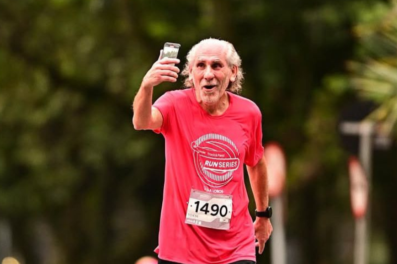 Homem idoso de camiseta rosa vibrante, correndo uma maratona e tirando uma selfie com o celular