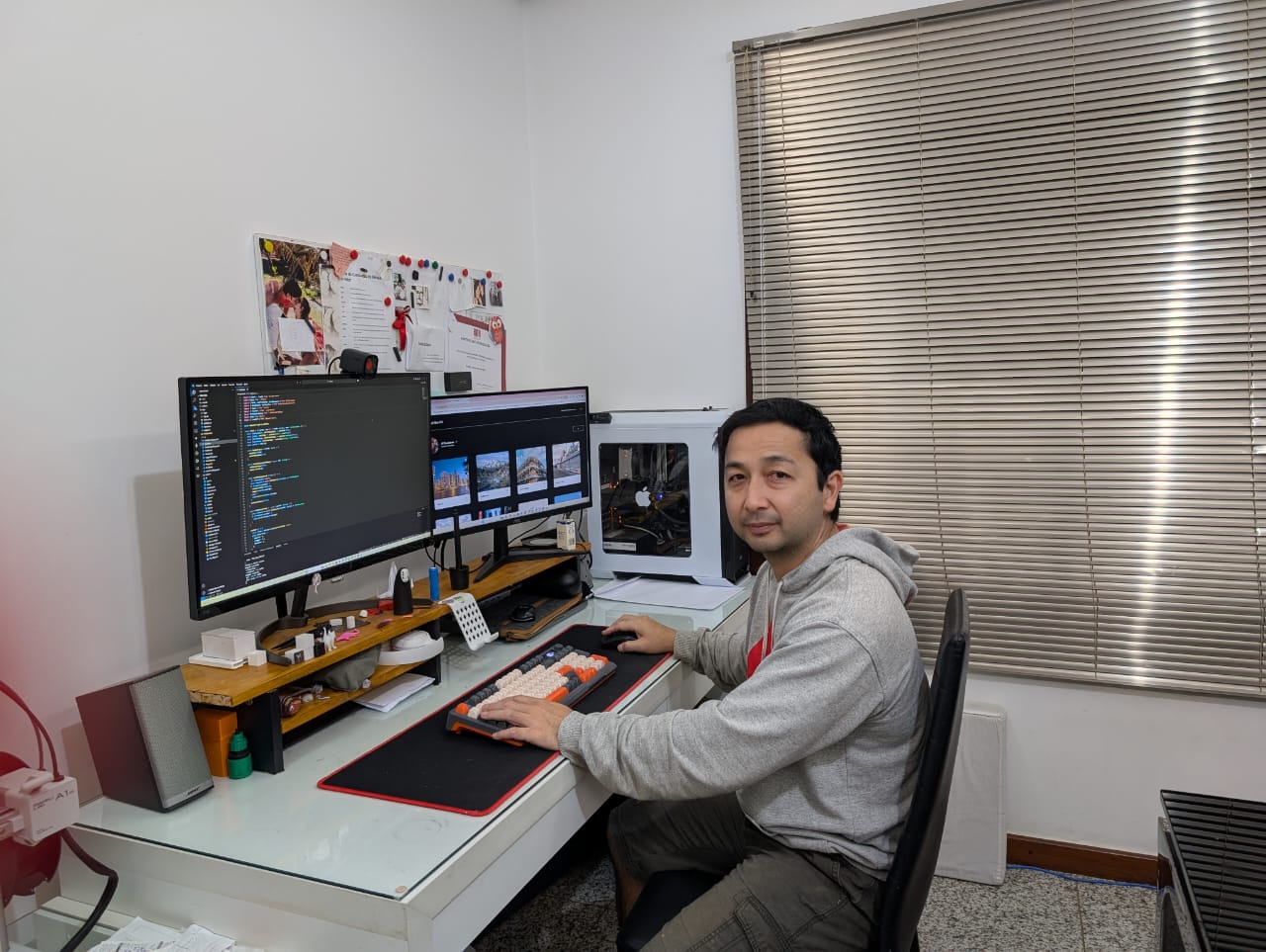 Alysson Tomizawa sentado em frente à messa de trabalho, onde estão dois monitores, olha para a câmera com a mão esquerda sobre o teclado