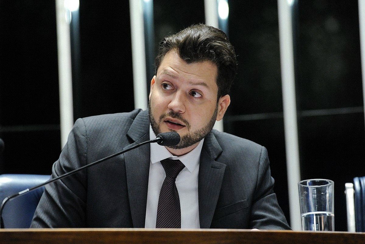 Daniel Nascimento em roupas sociais, sentado em uma mesa do plenário, com um copo d'água, enquanto fala ao microfone