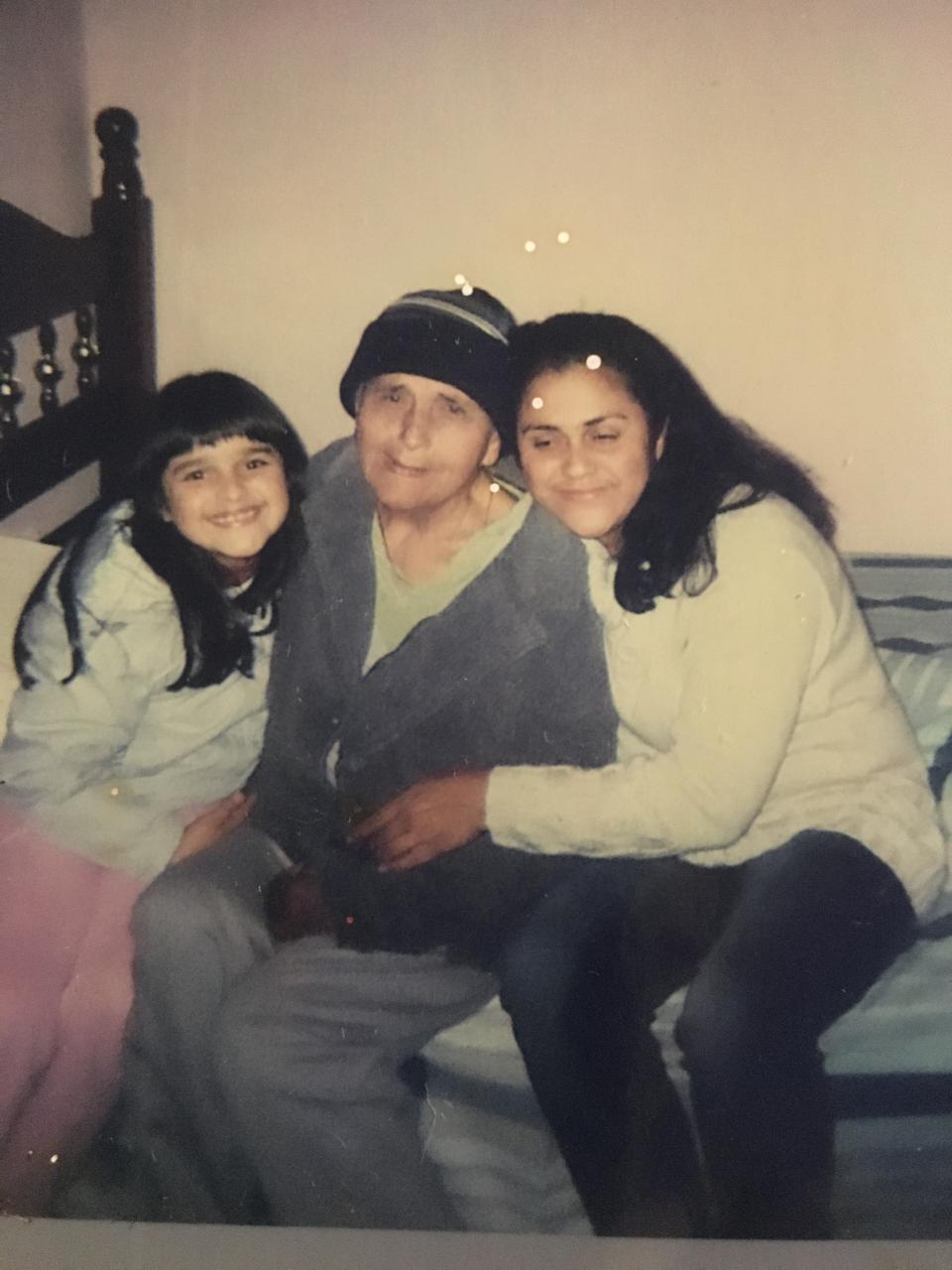 Idosa de gorro abraçada por criança e jovem em casa, representando o papel do cuidador familiar informal.