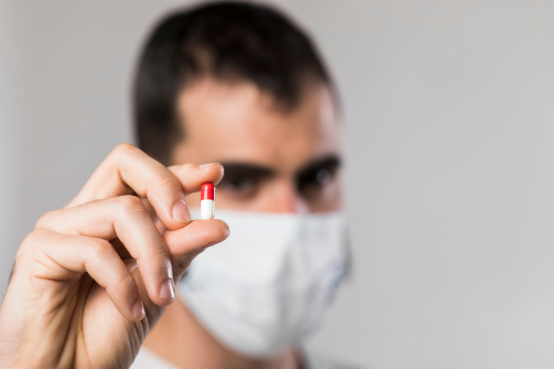 Homem de cabelos escuros e máscara segura uma pilula