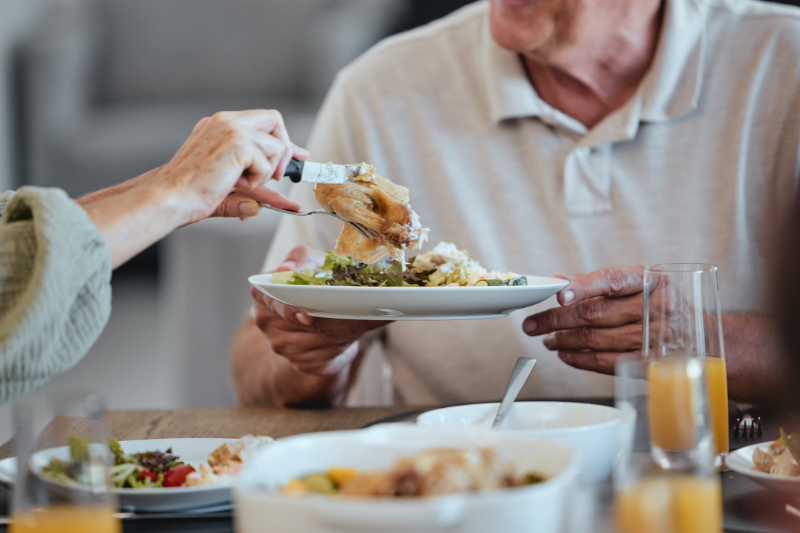 Homem idoso segura prato de comida recheado de legumes e uma pessoa o serve com pedaço de frango