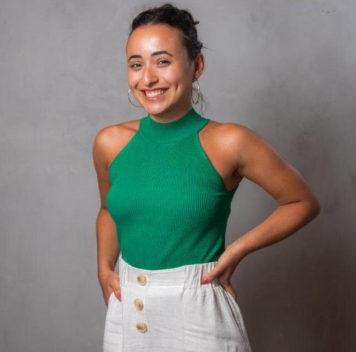 De pé, Larissa Carvalho sorri para a câmara, usando blusa verde e calça bege; cabelos pretos estão presos