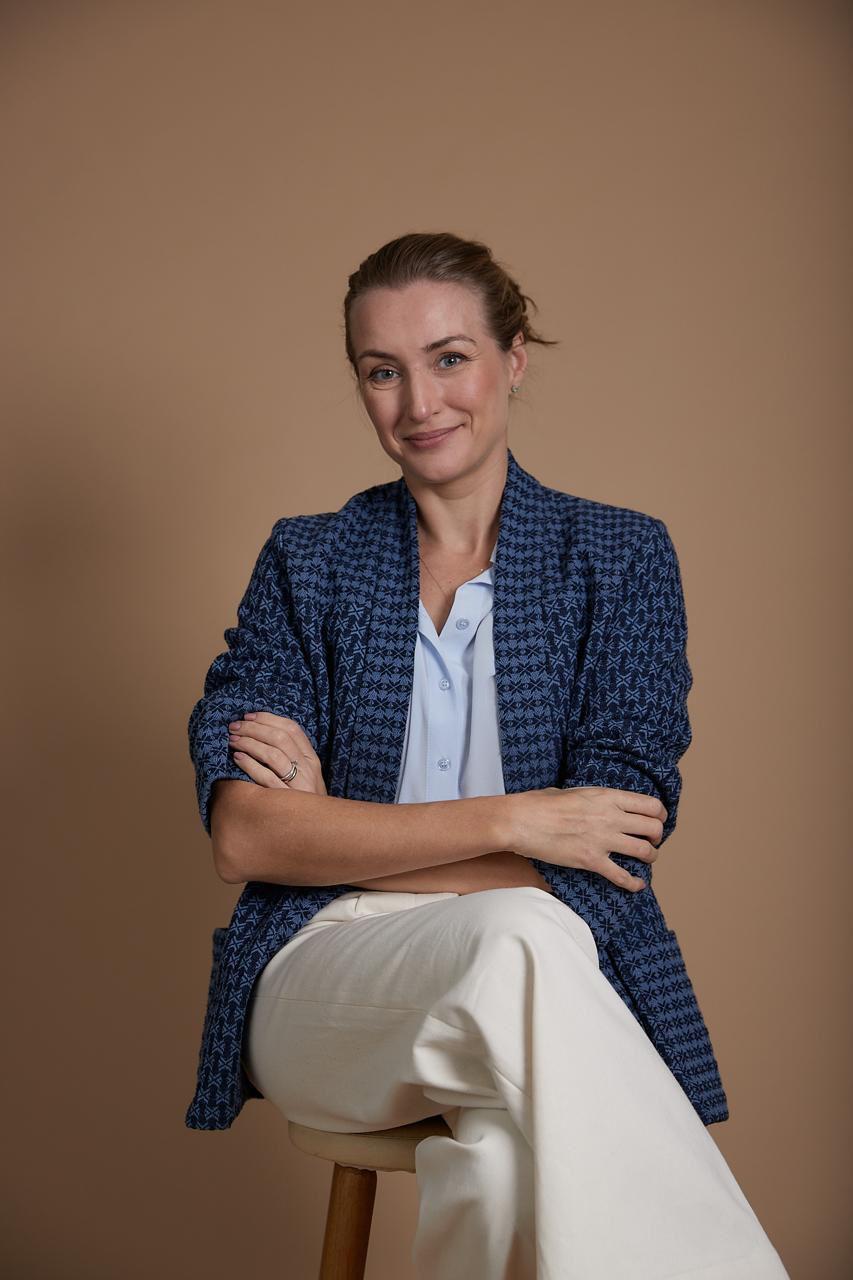 Milena Brentan sentada, de braços cruzados, cabelos pretos presos, usando blazer azul marinha e calça bege