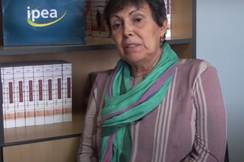 Pesquisadora ana amélia está sentada e olha para o lado enquanto fala, há uma estante com livros atrás dela