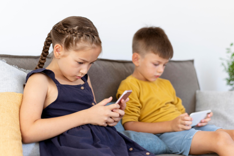 duas crianças, um menino e uma menina, sentados em um sofá mexendo no celular
