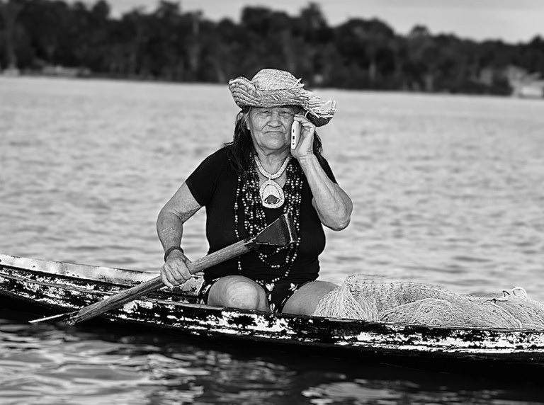 Foto em preto e branco. Uma mulher idosa fala ao celular enquanto conduz uma canoa em um grande rio