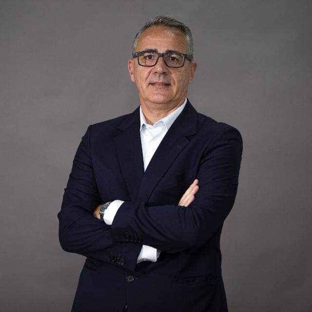 O professor Marcelo Treff, de pé, de óculos, braços cruzados, sobre paletó escuro, sob o qual veste uma camisa brancamisa