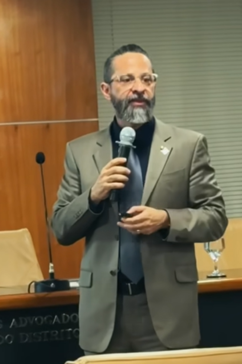 especialista em cibersegurança rodrigo fragola segura microfone, enquanto palestra para uma audiência. ele usa terno cinza, óculos e tem uma barba longa