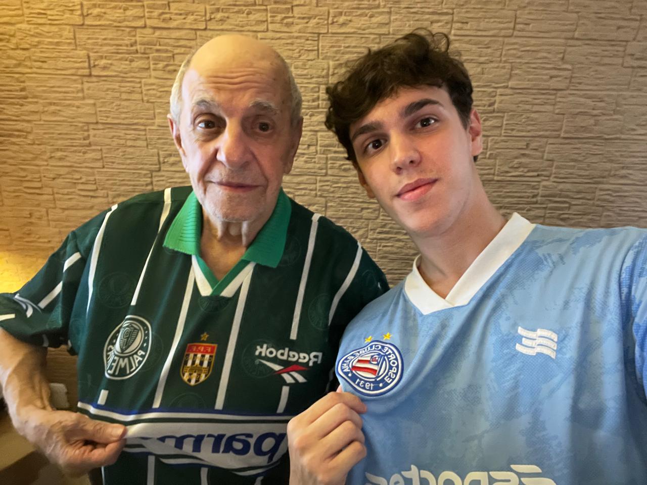 Adolpho, idoso com Alzheimer, e o neto Rafael tiram selfie mostrando suas camisas de time de futebol.
