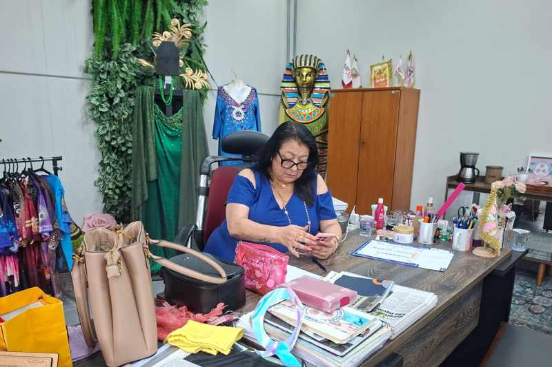 Angelina em seu escritório, sentada com vestido azul sentada mexendo no celular