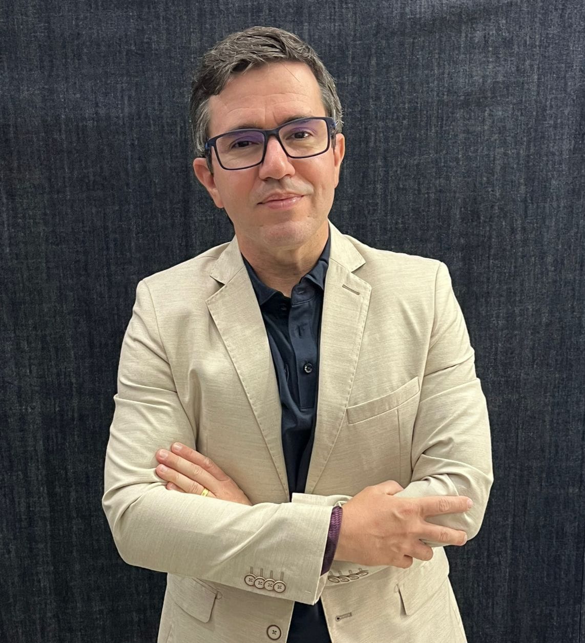 Gustavo Faria posa de pé, de blazer claro sobre uma camisa escura, com braços cruzados, usando óculos e esboçando um leve sorriso