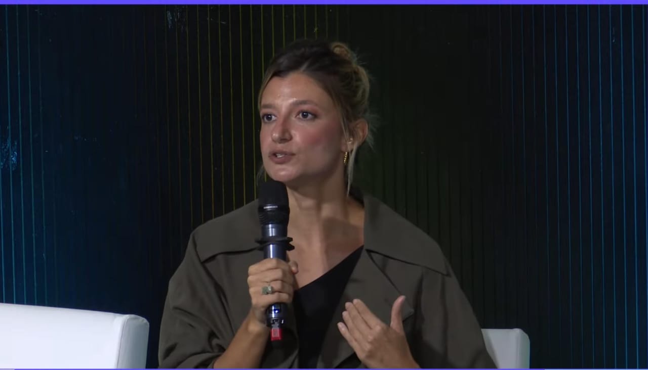 Marilia é uma mulher branca, está com os cabelos loiros presos e segura um microfone