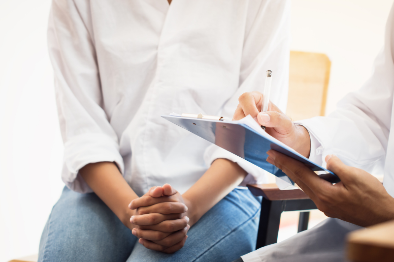Mulher branca sentada em um consultório com as mãos juntas enquanto médico anota sintomas