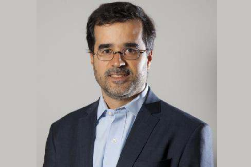 Caio Pereira é um homem branco, de meia idade com cabelos pretos, usa  óculos e está de terno azul