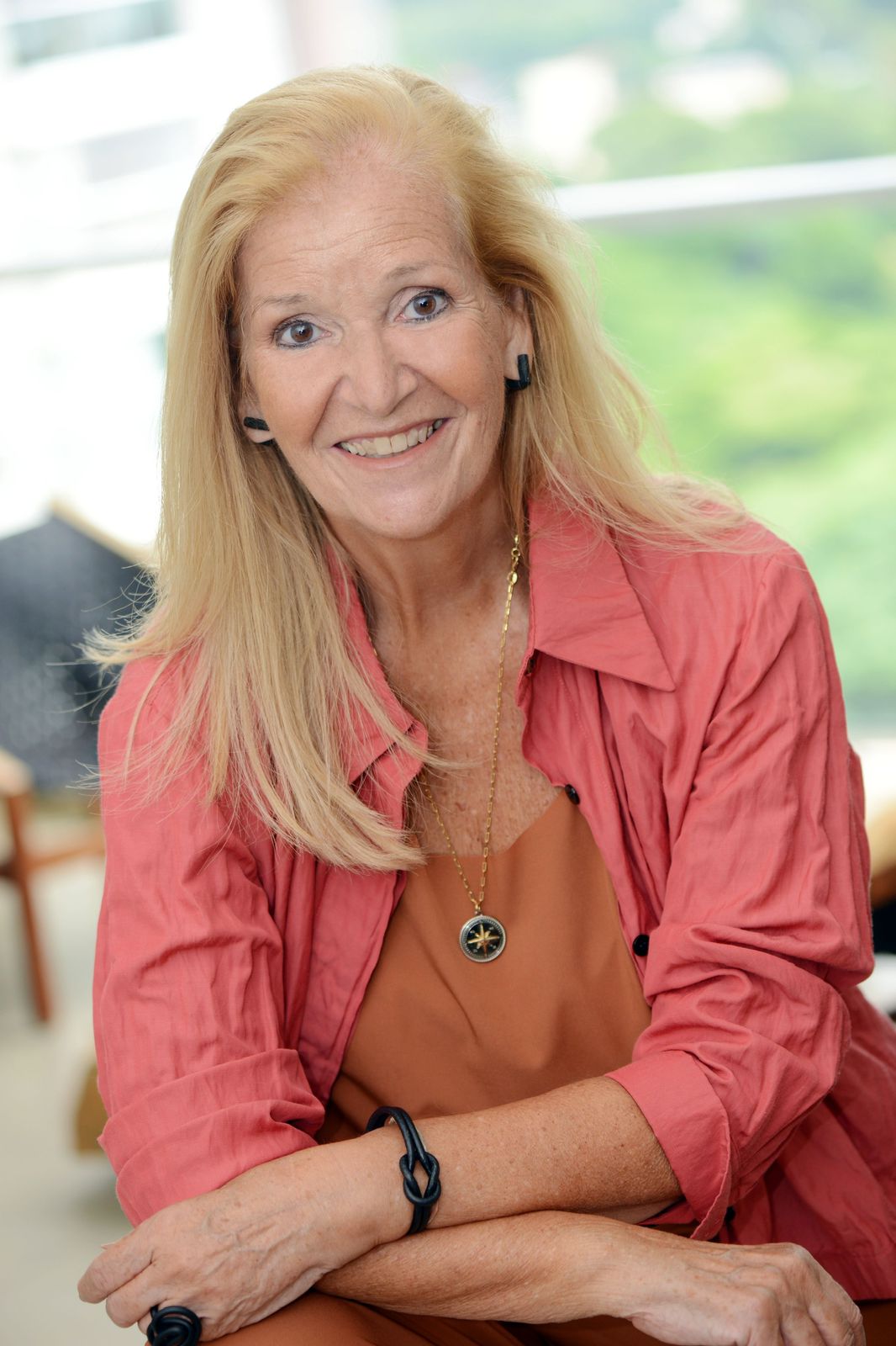 Chris Aché de cabelos longos à altura do peito, sentada sorrindo usando camisa rosa aberta sobre uma blusa ocre com uma corrente por cima dela