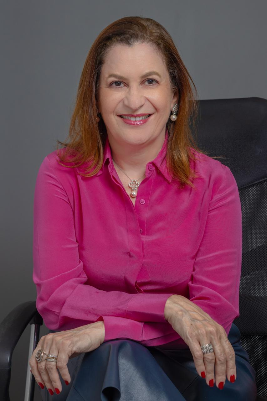 Elvira Presta sentada, sorrindo, tem cabelos à altura dos ombros; usa camisa cor de rosa 