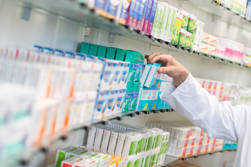 Braço com jaleco pega um remédio em prateleira com diversos medicamentos