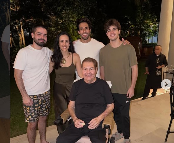 Faustão sentado em uma cadeira de rodas tendo atrás, em pé, os filhos e o genro