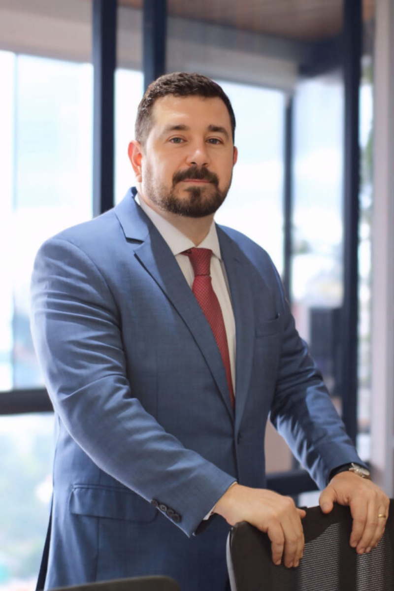 Leandro Petrin é um homem de meia idade de cabelos ralos e escuros, está de pé e segura uma cadeira. está vestido com um terno azul e gravata vermelha