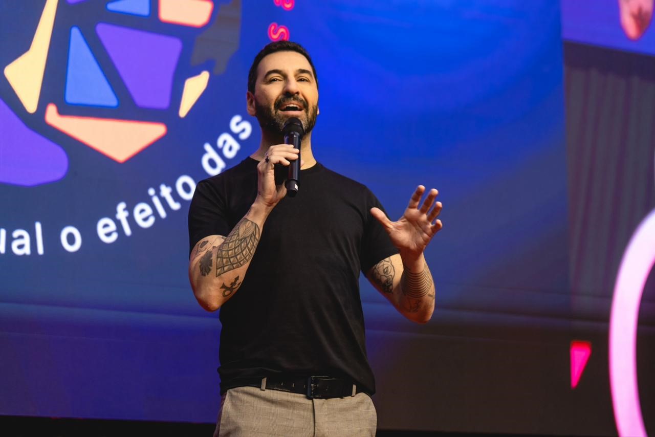 Marc Tawil fazendo uma palestra aparece de pé, com o microfone na mão de direita em frente a um painel azul