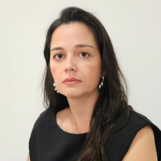 Priscila é uma mulher branca magra de cabelos compridos e escuros. Veste uma regata preta e olha séria para foto