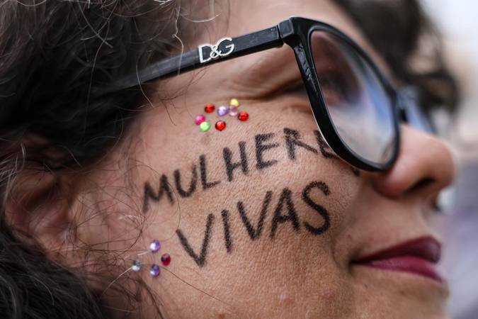 Mulher de óculos com a frase "MULHERES VIVAS" escrita no rosto, em protesto contra violência à mulher.