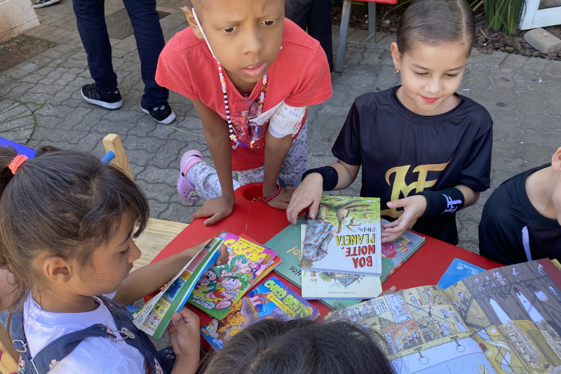 crianças, um menino e três menina sentados em uma mesa pequena escutam histórias das voluntárias do TUCCA