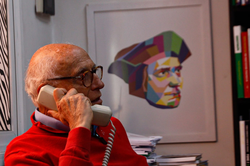 Eduardo Suplicy é um homem branco e idoso de cabelos brancos, veste um sueter vermelho e está sentado em uma cadeira enquanto fala no telefone