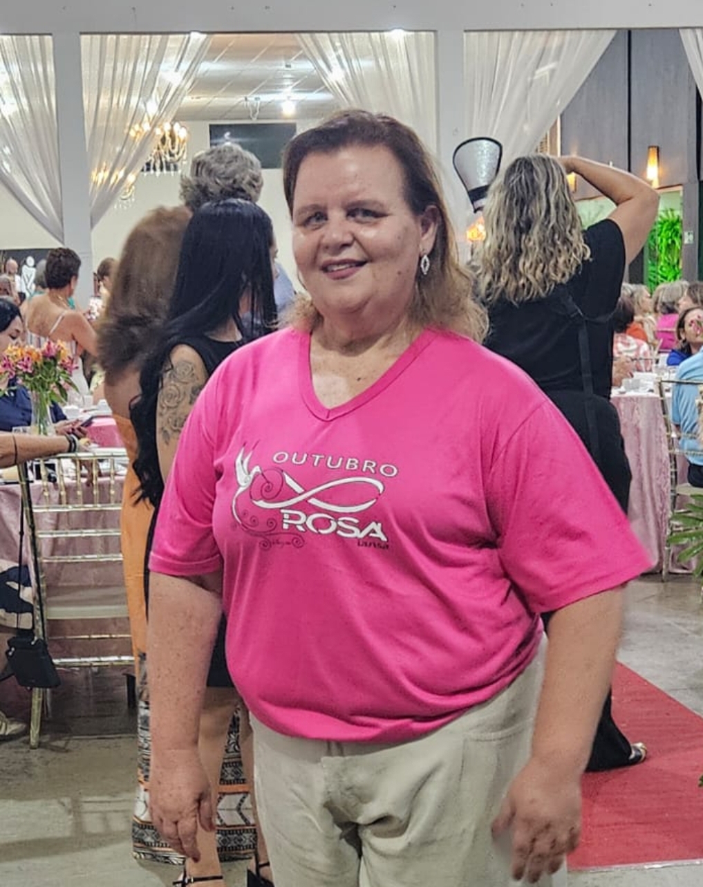 Márcia é uma mulher de meia idade que veste uma camiseta rosa. Ela tem rosto redondo e cabelo médio loiro