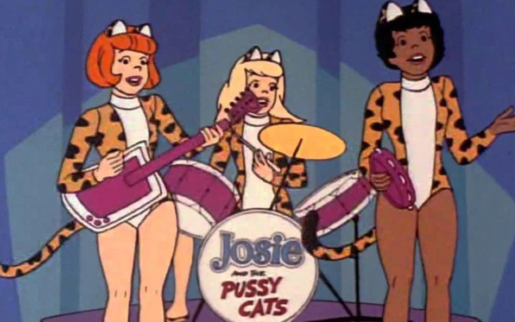 As três integrantes de Josie e as Gatinhas estão cantando em cima de um palco pequeno e usando maiô de oncinha