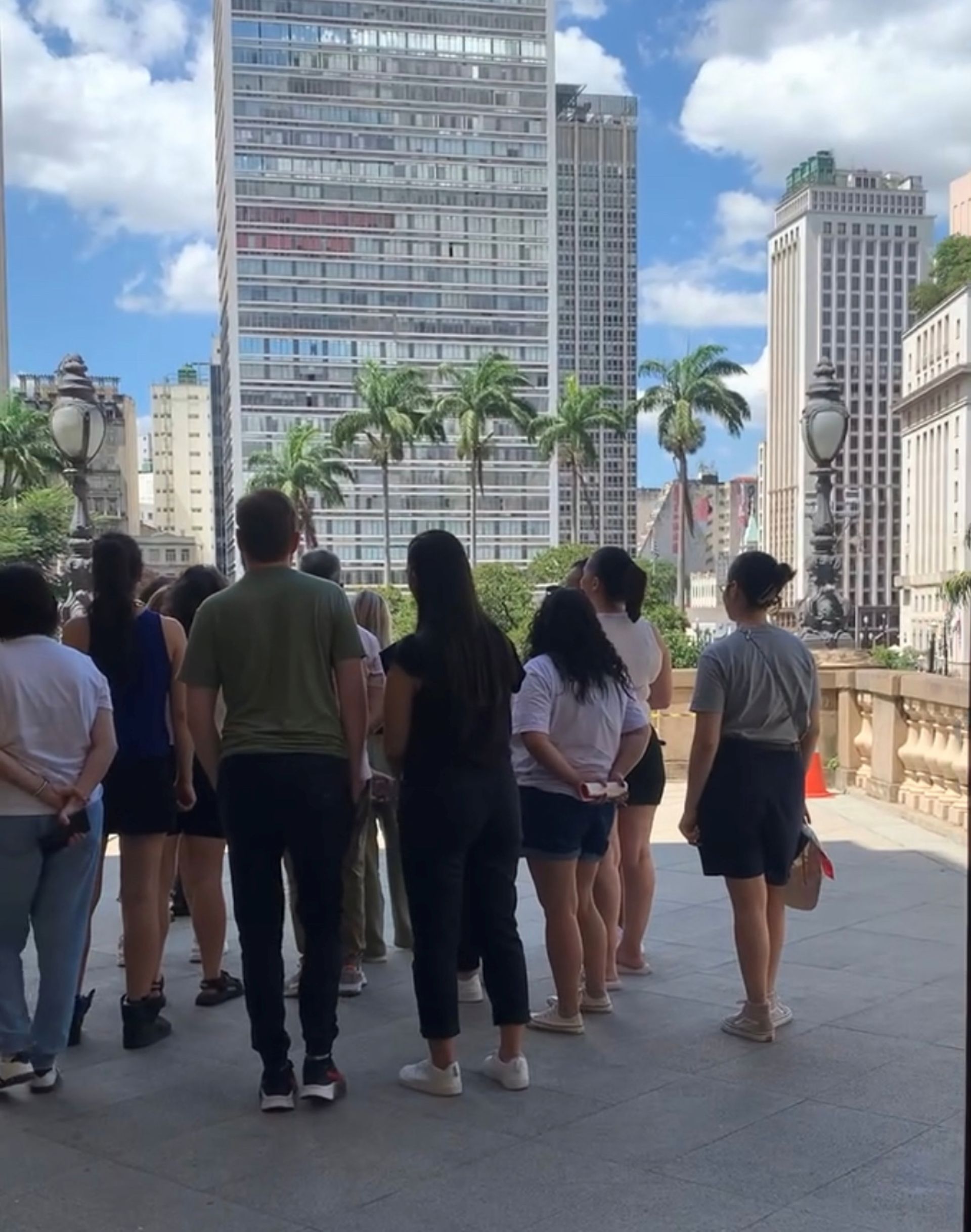 Visitantes estão na varanda, num dia de sol, ouvindo a educadora que conduziu o tour pelo Municipal