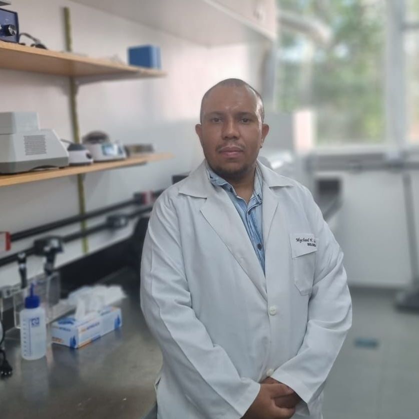 Mychael é um homem negro que veste um jaleco, está de braços cruzados e está em frente a uma estante de laboratório