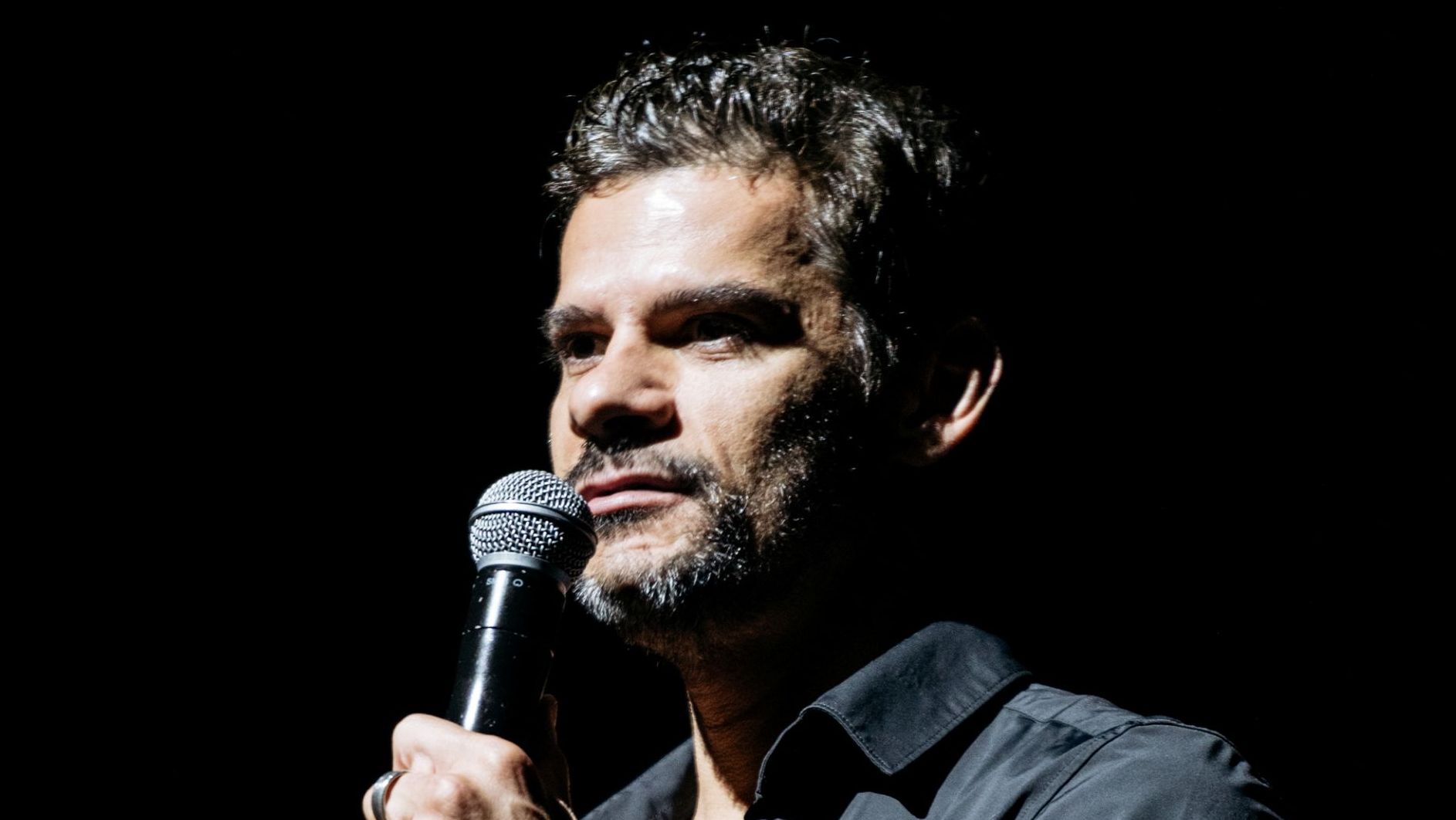 Thiago Soares está usando uma camisa preta e está segurando um micronfone