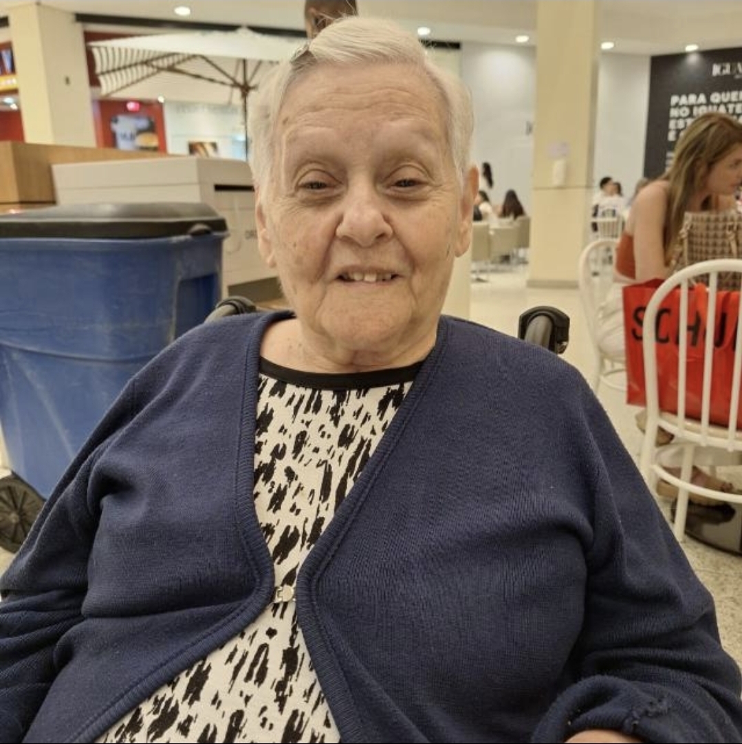 Marly é uma mulher idosa que está sentada, usa uma blusa branca e preta com cardigan azul