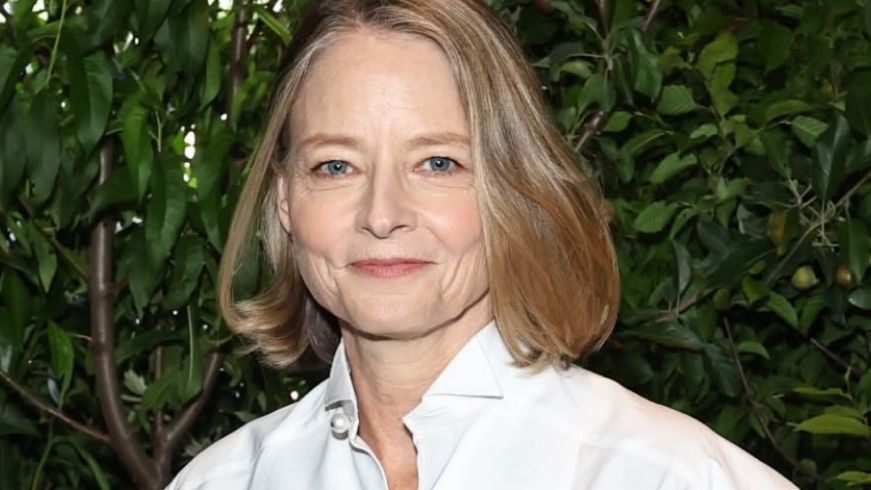 Jodie Foster usa camisa branca, cabelo loiro e sorri para a foto