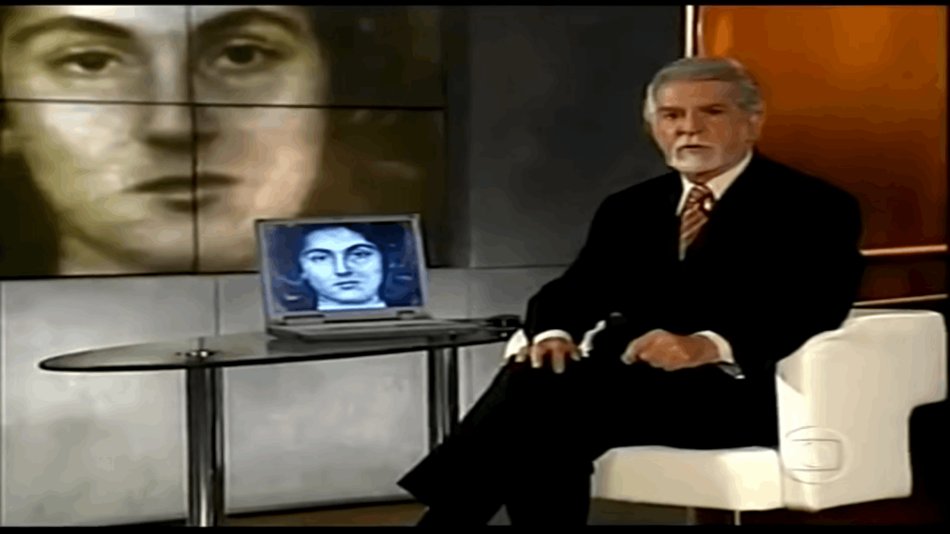 Apresentador do programa sentado, com um tablet com a foto da vítima, e uma grande imagem dela ao lado
