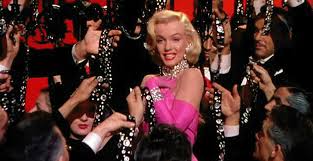 Marilyn Monroe usa vestido rosa e está cercada por mãos que oferecem a ela diamantes
