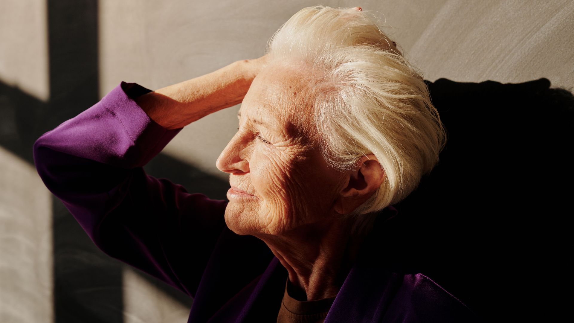 Mulher idosa de perfil, com cabelos brancos, iluminada pela luz do sol enquanto repousa a mão sobre a cabeça