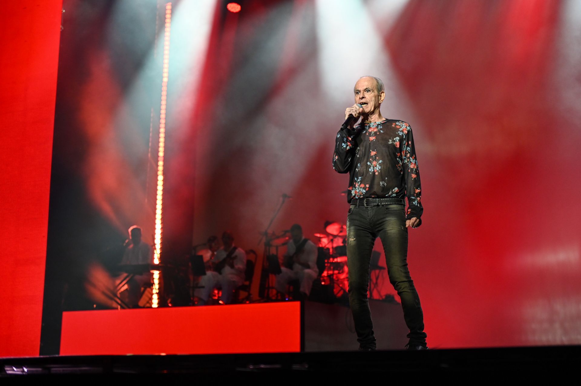 Ney Matogrosso usa roupa escura e está cantando em cima do palco, com o microfone na mão