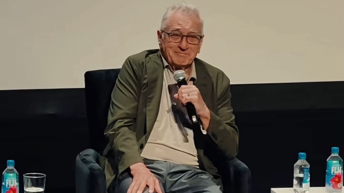 Robert De Niro está sentado em uma cadeira, e sorri para o público enquanto segura um microfone