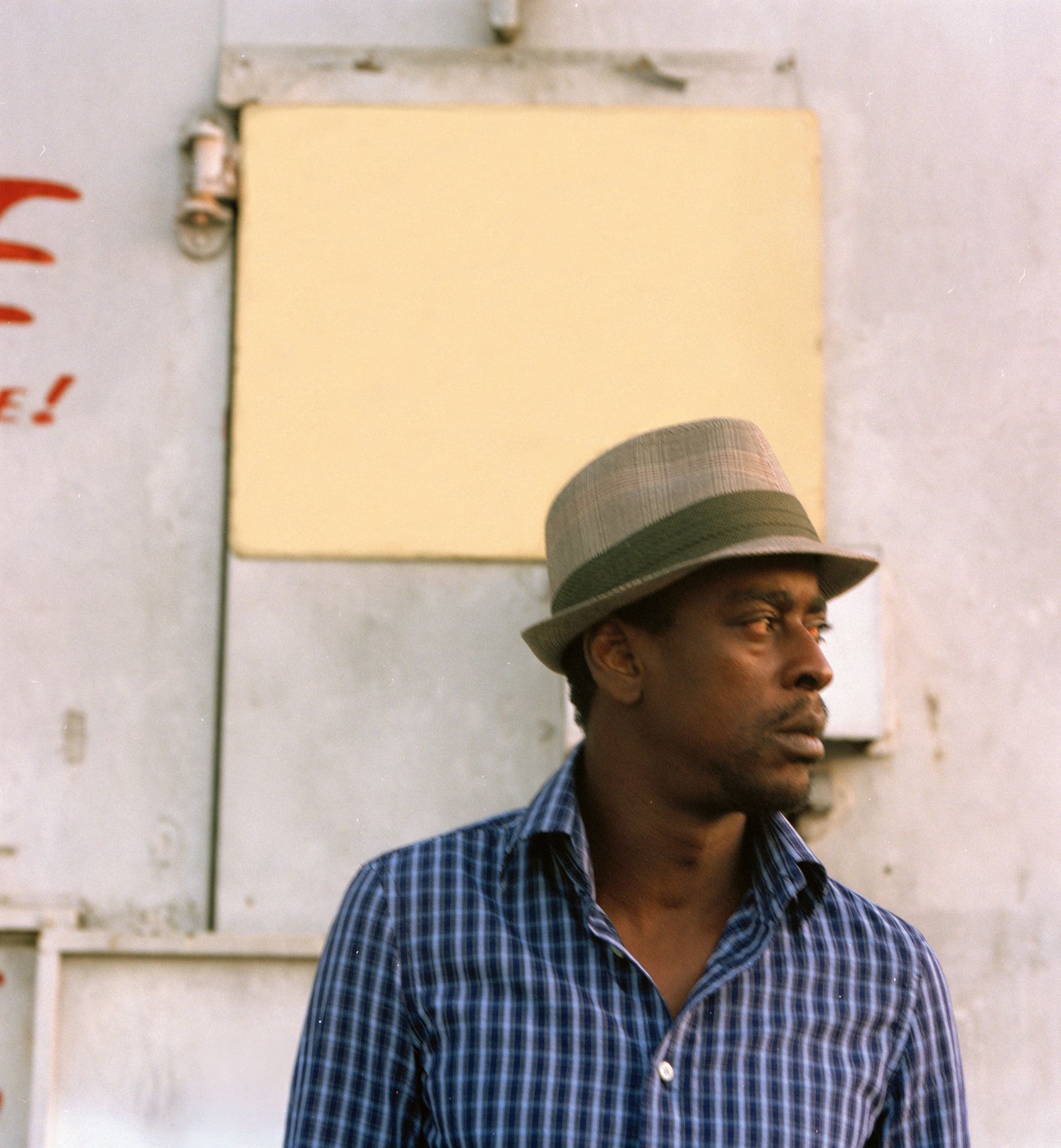 Seu Jorge usa camisa azul, chapéu e calça preta e está olhando para o lado