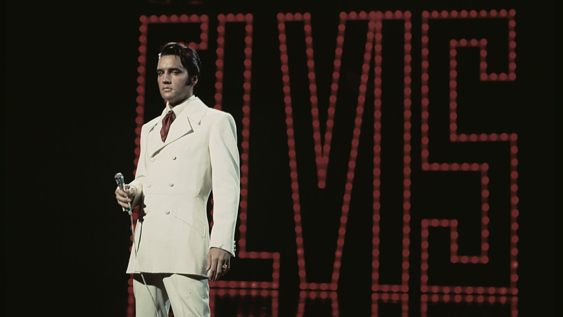 Elvis está usando terno e calça brancas, em frente a um letreiro com seu nome