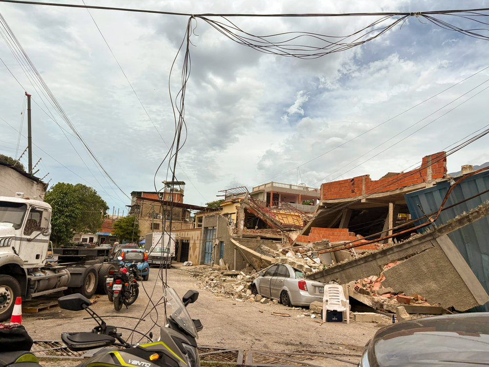Escombros de edifício desabado pelo terremoto bloqueiam rua com carros estacionados e fiação caída na Venezuela.