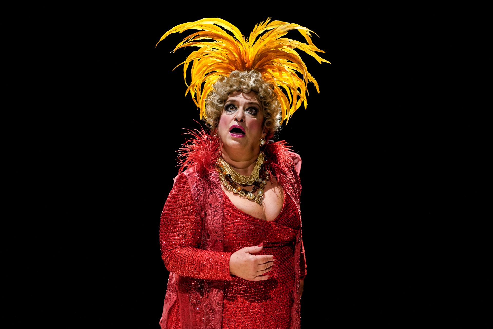 Grace Gianoukas está caracterizada como a atriz e comediante Dercy Gonçalves, com roupa vermelha e penas amarelas nos cabelos
