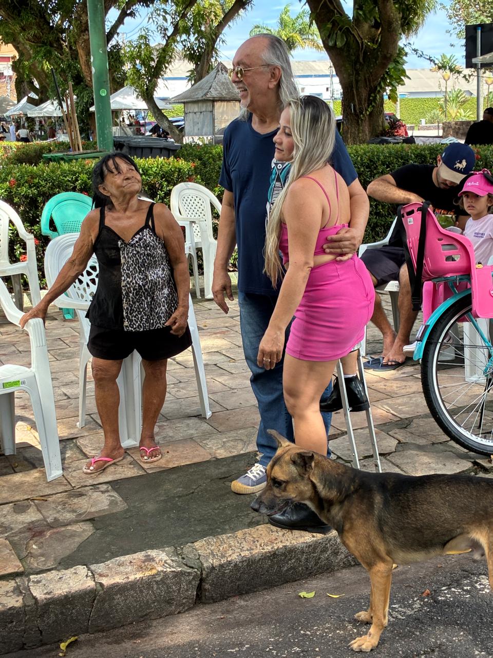 Cantor Nilson Chaves sorri abraçado a mulher de rosa em praça de Belém; um cão passa à frente.